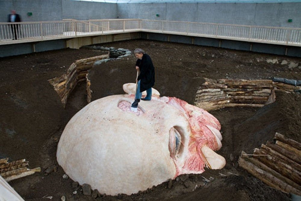 Inside a dirt pit a person digs into the brain of a giant human head.