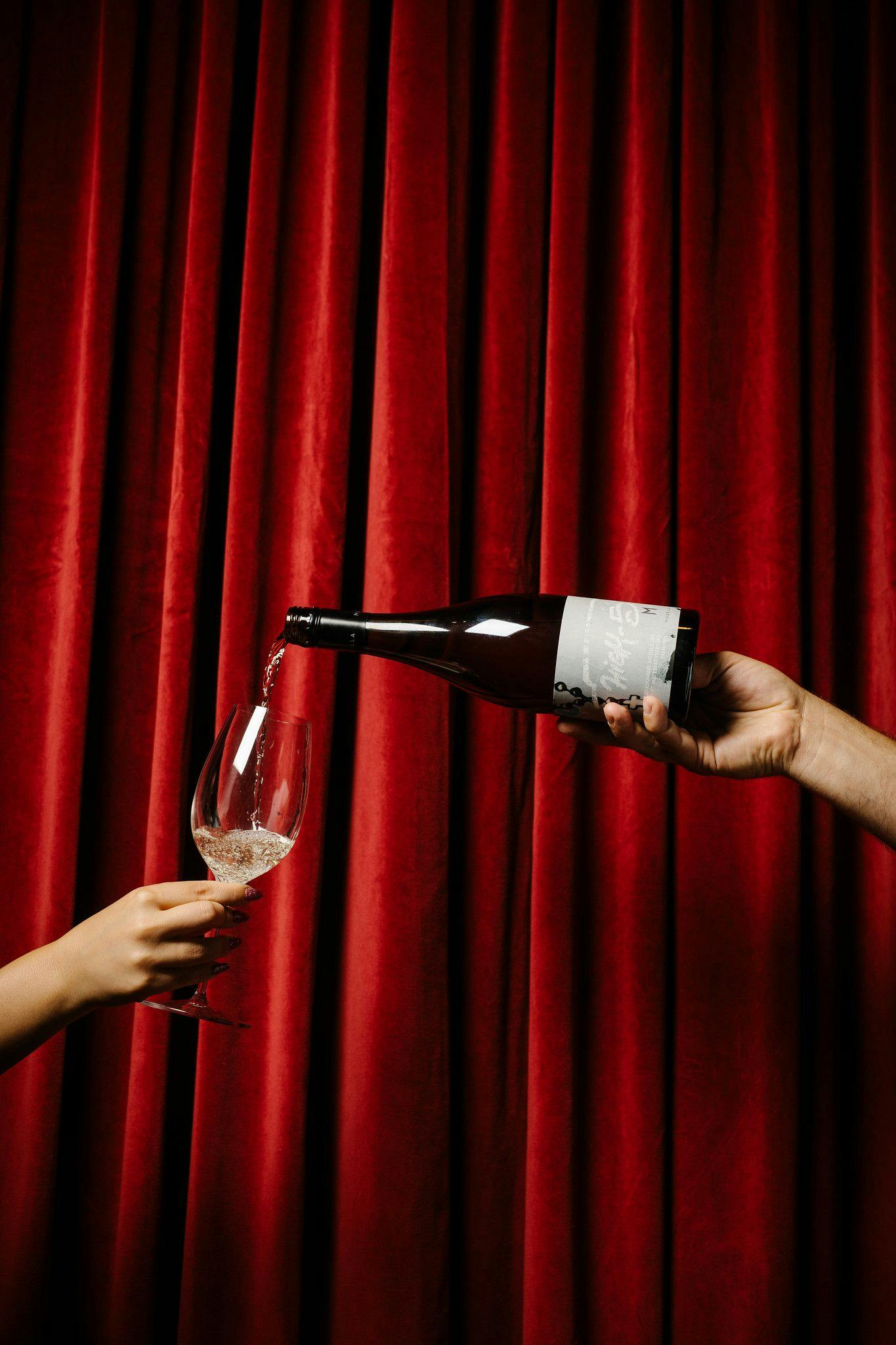 Hand of a waiter expertly pours a glass of bubbles in front of a luxurious red velvet curtain