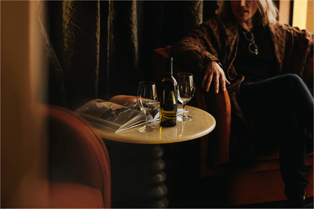 A person lounging in a chair next to a side table with two glasses, a bottle of wine and an open book