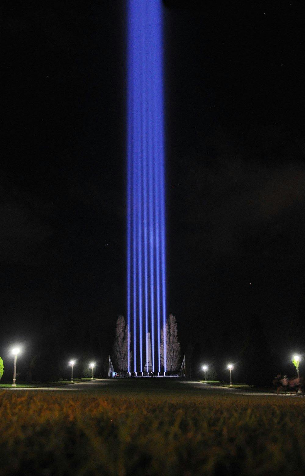 A series of blue lights are being beamed directly upwards creating a stream of light. Located in a park.