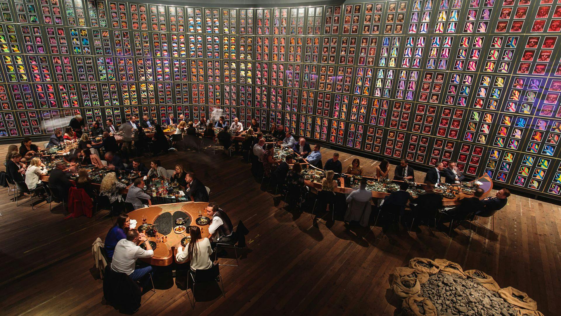 A long table of diners shaped like a snake under a vast wall of images that come together to form the image of a snake