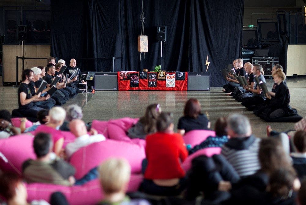 Two rows of people chanting from books infront of an altar of Amplifiers. Audience watches in the foreground.