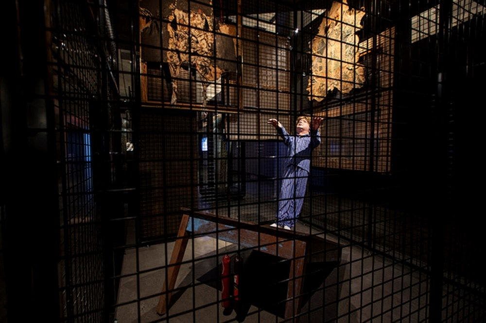 A black cage surrounds a figure of a sleepwalking man boy on a table with salamis hanging.